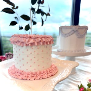 May include: A cylindrical cake with white frosting and small pink polka dots. The cake features a ruffled pink frosting border at the base and a matching ruffled pink frosting trim along the top edge. A second white cake with blue trim is visible.