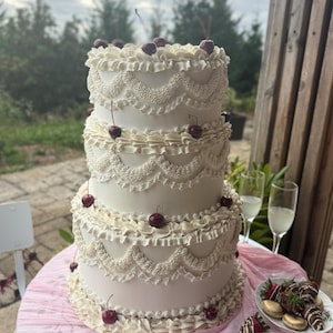 May include: A three-tiered white cake decorated with intricate piped buttercream ruffles and swags, topped and accented with fresh red cherries. The cake sits on a table draped with a light pink fabric, accompanied by a plate of chocolate-covered strawberries.