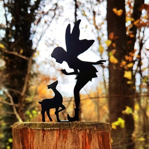 May include: A black metal silhouette of a fairy with wings outstretched, holding out a hand to a small deer silhouette, both mounted on a wooden post in a woodland setting.