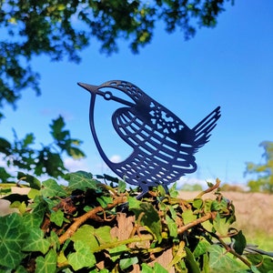 May include: A black metal silhouette of a bird with intricate details, perched on a wooden post covered in green ivy.