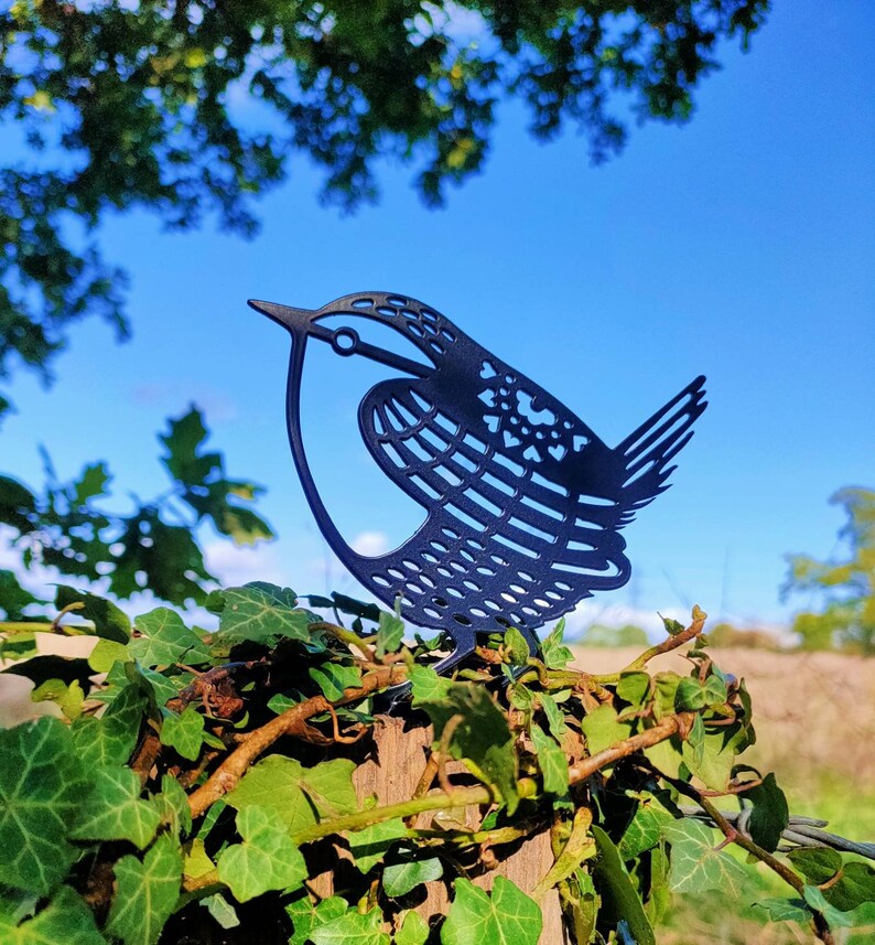 May include: A black metal silhouette of a bird with intricate details, perched on a wooden post covered in green ivy.