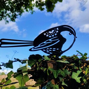 May include: Black metal silhouette of a bird perched on a wooden post with green ivy growing around it. The bird is facing right and has a detailed design.