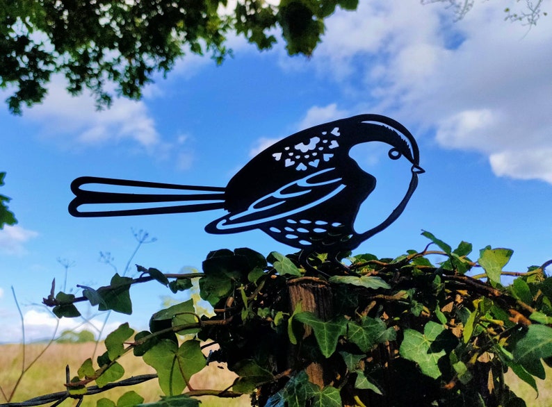 May include: Black metal silhouette of a bird perched on a wooden post with green ivy growing around it. The bird is facing right and has a detailed design.