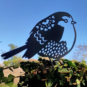 May include: Black metal silhouette of a bird with intricate cut-out designs. The bird is perched on a bed of green foliage.