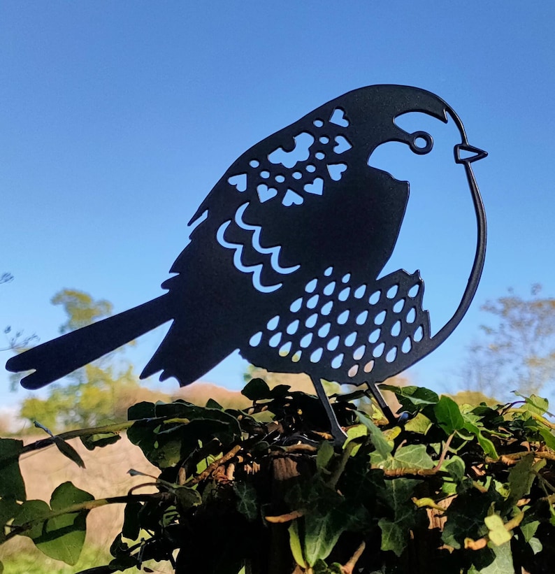 May include: Black metal silhouette of a bird with intricate cut-out designs. The bird is perched on a bed of green foliage.