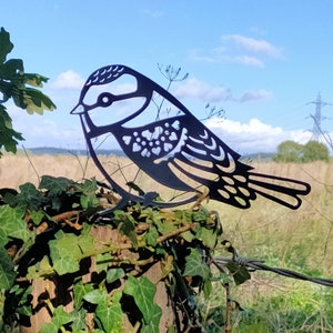 May include: Black metal silhouette of a bird with intricate details, perched on a wooden post covered in green ivy.