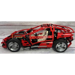 May include: A red sports car model constructed from interlocking plastic building blocks. The vehicle features an open-frame chassis, visible white interior seating, a steering wheel, and four black wheels with silver-coloured rims. The structure is composed of red beams and connectors, showcasing a detailed mechanical design with exposed engine components and a roll cage frame.