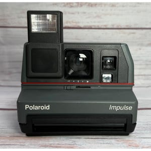 Può includere: Una fotocamera istantanea Polaroid Impulse grigia con flash a comparsa. La fotocamera ha un obiettivo nero e una linea di accento rossa. La parola "Polaroid" è stampata sulla parte anteriore, con "Impulse" sul lato destro.