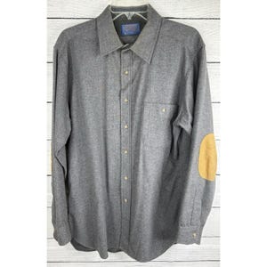 Puede incluir: Una camisa gris abotonada con mangas largas, un bolsillo en el pecho y coderas color canela. La camisa tiene cuello y está colgada de una percha. La camisa está hecha de un material suave, similar a la lana.