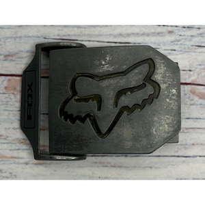 May include: A dark gray metal belt buckle featuring a distressed finish and an embossed Fox head logo in the center. The rectangular frame includes the word FOX vertically debossed on the left side, with a hinged mechanism for attachment.