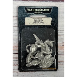 Puede incluir: Una figura miniatura Tyranid Tyrant Guard de color plateado de Warhammer 40,000, sellada en su empaque blíster negro original. El empaque presenta el logotipo de Warhammer 40,000 en la parte superior y una etiqueta con el nombre del producto.