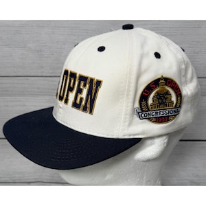 Op de afbeelding: Een witte baseball cap met een marineblauwe klep. De cap heeft het woord "OPEN" in marineblauw en goud. Een cirkelvormige patch aan de zijkant leest "U.S. OPEN CONGRESSIONAL 1997".