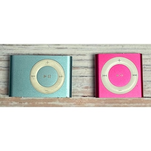 May include: Two rectangular digital music players, one in light blue and one in bright pink. Each device features a circular white control pad in the centre with symbols for play, pause, skip forward, skip backward, volume up, and volume down.