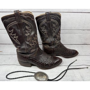 May include: A pair of brown leather cowboy boots with decorative stitching and a snakeskin pattern on the lower part. A black bolo tie with a silver-colored oval clasp is also present.