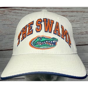 Peut inclure: Une casquette de baseball blanc cassé avec le texte brodé THE SWAMP en orange avec un contour bleu foncé. Sous le texte se trouve un graphique brodé représentant une tête d'alligator verte avec un contour orange. Bordure bleu foncé.