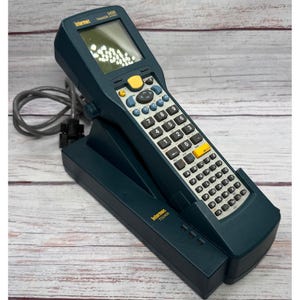 Pode incluir: Um computador portátil Intermec Trakker 2420 azul-escuro acoplado numa base Intermec TD2400. O dispositivo possui um ecrã, um botão de energia amarelo, um pad direcional, teclado numérico e teclado QWERTY completo com um cabo cinzento.