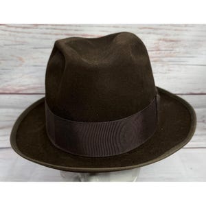 Puede incluir: Un sombrero fedora de color marrón oscuro que presenta una copa estructurada con un pliegue central y un ala ancha. El sombrero está acentuado con una cinta de grosgrain marrón oscuro a juego que rodea la base de la copa, uniforme.