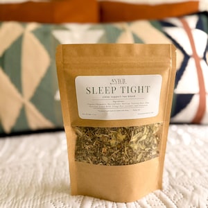 May include: A brown paper bag of "Sleep Tight" sleep support tea blend. The label includes the brand name "NWB" and a list of ingredients. The bag contains a mix of dried herbs and is set against a blurred background of pillows and a white bedspread. Net weight: 2 oz.