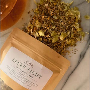 May include: A brown paper bag of "SLEEP TIGHT" sleep support tea blend with a pile of dried herbs and a glass of tea. The bag's label lists ingredients like chamomile and lavender. The text "Nourish with Brit" is also visible.