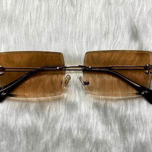 May include: Rimless sunglasses with rectangular brown tinted lenses and a gold-colored metal frame. The temples are black and have decorative accents. The glasses are set against a white, textured background.