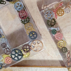 Pode incluir: Dois porta-retratos de resina transparente com uma borda decorativa de engrenagens douradas, prateadas e acobreadas. Os porta-retratos são retangulares e têm uma abertura dupla.