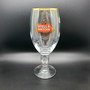 Może przedstawiać: Szklanka do piwa Stella Artois wykonana z przezroczystego szkła z pozłacanym rantem. Szklanka ma nóżkę i logo Stella Artois.