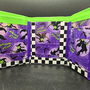 Può includere: Un portafoglio in nastro adesivo viola e verde con un design di strega e luna. Il portafoglio ha più tasche e un motivo a scacchi all'esterno.