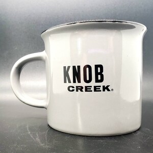 May include: A white ceramic mug with a black rim and handle. The mug has the words "Knob Creek" printed in black on the front.
