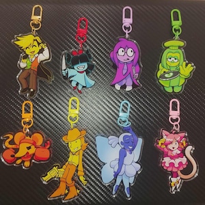 Op de afbeelding: Acht acryl sleutelhangers met diverse cartoonfiguren in levendige kleuren, elk bevestigd aan een bijpassende plastic karabijnhaak. De ontwerpen omvatten een figuur met geel haar, een blauw personage met een mes, een paars figuur, een groen personage, een oranje muis, een gele cowboy, een blauwe fee en een roze katachtig personage.