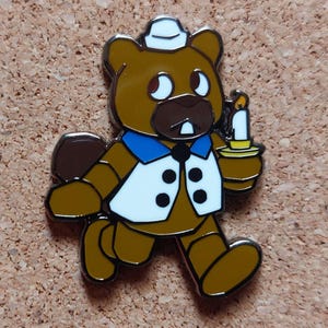 Pode incluir: Um alfinete de esmalte de urso marrom com colete branco, gola azul e um pequeno chap&eacute;u branco. O urso est&aacute; segurando uma vela com uma chama amarela. O alfinete tem um contorno prateado e est&aacute; em uma superf&iacute;cie de corti&ccedil;a.
