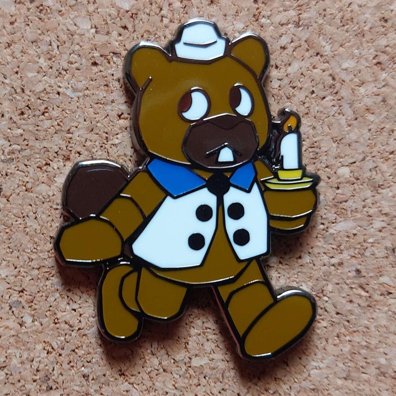 Pode incluir: Um alfinete de esmalte de urso marrom com colete branco, gola azul e um pequeno chap&eacute;u branco. O urso est&aacute; segurando uma vela com uma chama amarela. O alfinete tem um contorno prateado e est&aacute; em uma superf&iacute;cie de corti&ccedil;a.