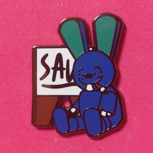May include: A blue bunny pin with green ears and a white sign that says "SAU". The bunny is holding a cigarette and is sitting down. The pin has a gold outline and is on a pink background.