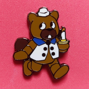 Pode incluir: Um broche de esmalte de urso marrom com colete branco, gola azul e chap&eacute;u branco. O urso est&aacute; segurando uma vela em um casti&ccedil;al dourado. O broche est&aacute; em um fundo rosa.