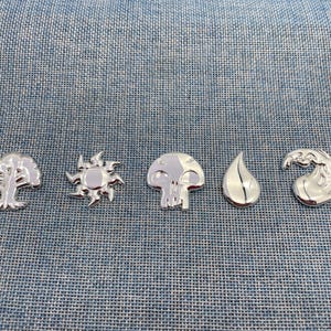 Op de afbeelding: Een set van vijf zilverkleurige metalen pins met verschillende vormen: een boom, een zon met puntige stralen, een schedel, een druppel en een kolkende golf. Elke pin heeft een gepolijst, reflecterend oppervlak en een gladde afwerking.