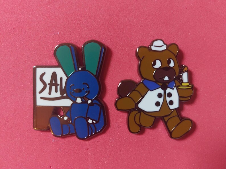 Pode incluir: Dois pinos de esmalte sobre um fundo rosa. Um pino apresenta um coelho azul com orelhas verdes e um sinal que diz "SAV". O outro pino &eacute; um urso marrom segurando uma vela.