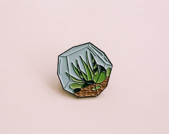 Puede incluir: Una planta suculenta verde dentro de un terrario de vidrio geométrico con una base marrón. El terrario es un pin con un respaldo plateado.