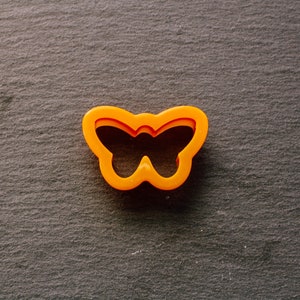 Puede incluir: Cortador de galletas de plástico naranja en forma de mariposa sobre un fondo gris.