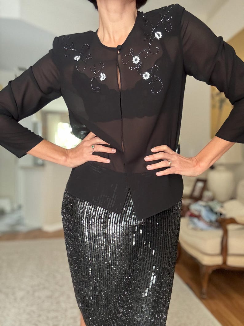 May include: A black sheer blouse with floral embroidery and a matching black sequined pencil skirt. The blouse has long sleeves and a button closure. The skirt is knee-length and covered in shimmering sequins.