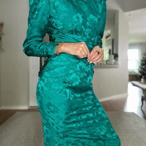 May include: A teal blue dress with a floral pattern. The dress has long sleeves and a fitted silhouette. It features a round neckline and a gold chain necklace.