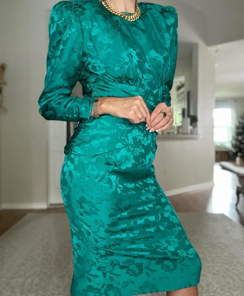 May include: A teal blue dress with a floral pattern. The dress has long sleeves and a fitted silhouette. It features a round neckline and a gold chain necklace.