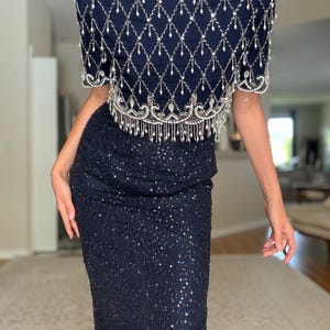 May include: A navy blue two-piece formal dress. The top is embellished with silver beading in a diamond pattern, with a beaded fringe along the bottom. The skirt is a matching navy blue, covered in sequins. The dress is a vintage style.