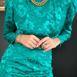 May include: A teal blue dress with a floral pattern and long sleeves. The dress has a fitted bodice and a slightly flared skirt. The model is wearing a gold chain necklace and a silver bracelet.