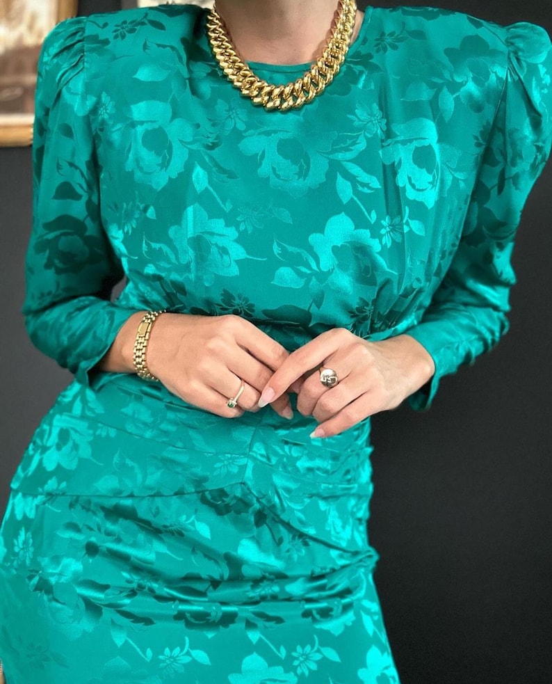May include: A teal blue dress with a floral pattern and long sleeves. The dress has a fitted bodice and a slightly flared skirt. The model is wearing a gold chain necklace and a silver bracelet.