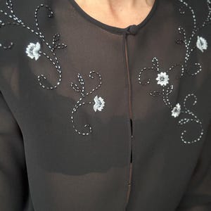 May include: Black sheer blouse with white floral embroidery. The blouse features a round neckline and a single button closure. The floral embroidery is detailed with white thread and small white flower accents.