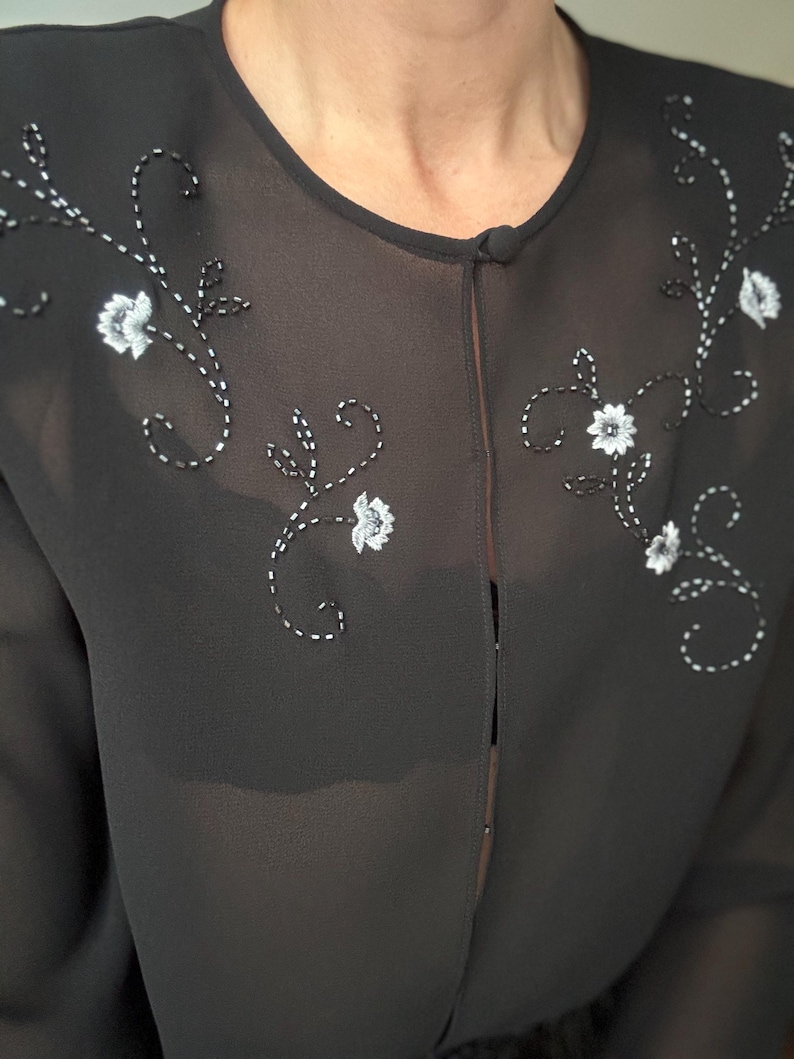 May include: Black sheer blouse with white floral embroidery. The blouse features a round neckline and a single button closure. The floral embroidery is detailed with white thread and small white flower accents.