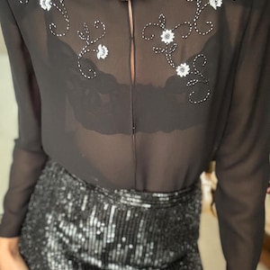 May include: Black sheer blouse with floral embroidery and a sequined skirt. The blouse features white floral embroidery and a keyhole neckline. The skirt is covered in black sequins, reflecting light. A silver beaded necklace is also visible.