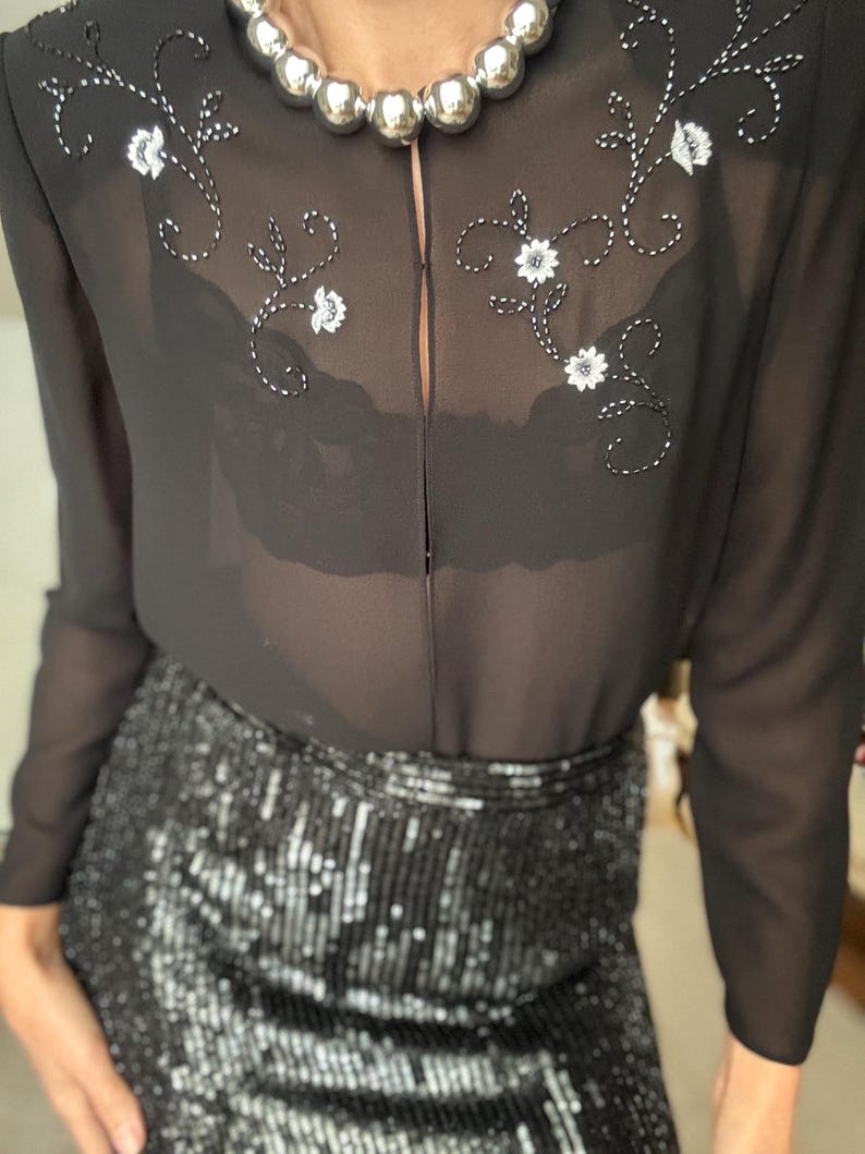 May include: Black sheer blouse with floral embroidery and a sequined skirt. The blouse features white floral embroidery and a keyhole neckline. The skirt is covered in black sequins, reflecting light. A silver beaded necklace is also visible.