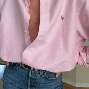 Puede incluir: Una camisa rosa claro con botones y un pequeño logo bordado en el pecho. Se combina con vaqueros azules y un cinturón negro con hebilla dorada. La camisa está desabrochada en la parte superior.
