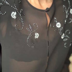 May include: A sheer black blouse with floral embroidery. The blouse features a round neckline and a single button closure. Delicate white and silver floral embroidery adorns the shoulders and front of the blouse, creating a subtle, elegant design.