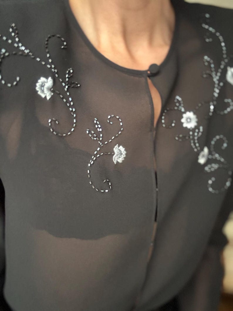 May include: A sheer black blouse with floral embroidery. The blouse features a round neckline and a single button closure. Delicate white and silver floral embroidery adorns the shoulders and front of the blouse, creating a subtle, elegant design.